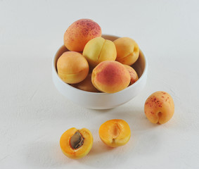 Ripe apricots in a white porcelain bowl. Several apricot fruits lie next to the bowl. One apricot is broken in half. Close-up. White background.