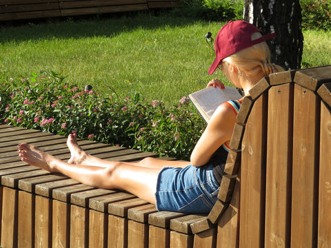 Barefoot Blonde Girl In Baseball Cap And Denim Shorts Reading A Book Sitting On A Wooden Bench. College Education, Student Preparing To The University, Reading Girl, Tanned Legs, Summer Fashion