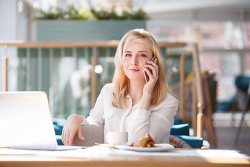 Cute young woman communicates on the phone with friends sitting at a table in a cafe on sunny day in cozy atmosphere.