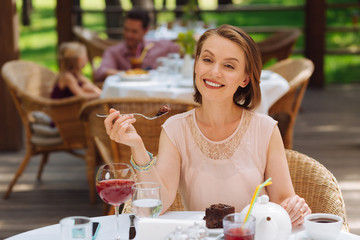 Joyful time. Beautiful smiling woman enjoying tasty cake and glass of wine while sitting outside on summer terrace