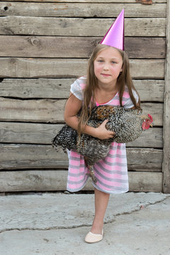Young girl in pointed magic hat holds motley chicken in her hands and poses in front of camera in village courtyard. The concept of healthy lifestyle in nature, love of the world, respect for nature