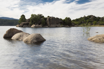 Vista de un lago con rocas en primer término y más rocas con árboles en el fondo