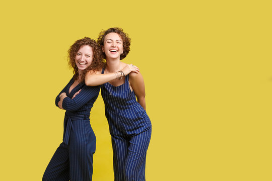 Studio Shot Of Two Caucasian Sisters With Same Curly Hair, Wearing Similar Blue Stripped Overall, Smiling Happily, Looking At Camera, Posing At Yellow Blank Wall, Standing To Each Other Closely