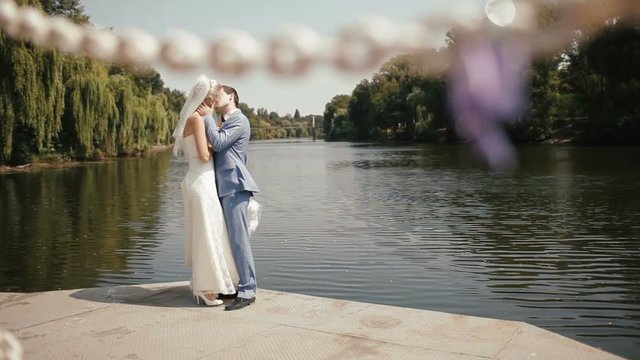Close-up view of the love lock wrapped in the pupple ribbon at the background of the beautiful kissing couple of newlyweds near the riber bank.