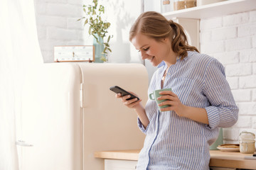 Fototapeta na wymiar Young woman using smartphone leaning at kitchen table with coffee mug and organizer in a modern home. Smiling woman reading phone message. Brunette happy girl typing a text message.