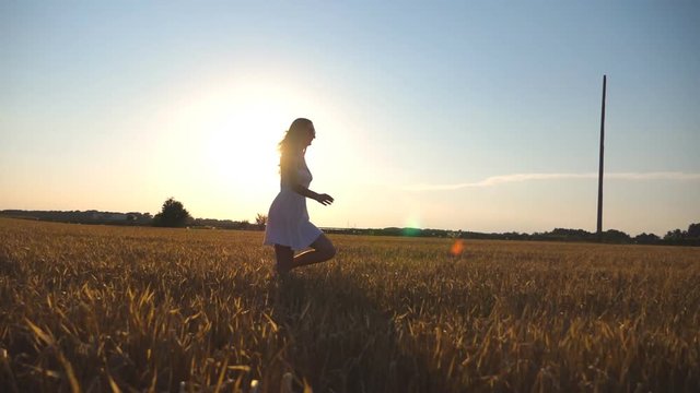 Young girl is running along wheat field under blue sky at sunset. Woman is jogging at the meadow. Sun shine at background. Side view Slow motion