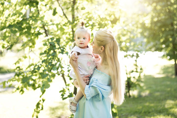 Fototapeta premium Young beautiful blonde mother with her baby girl laughing together and playing in green park outdoors at sunny day