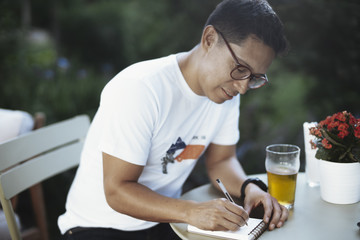 Young spectacled man drinking cold beer and writing some notes.