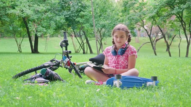 Beautiful young girl reads in the park on the grass next to her bicycle and skateboard