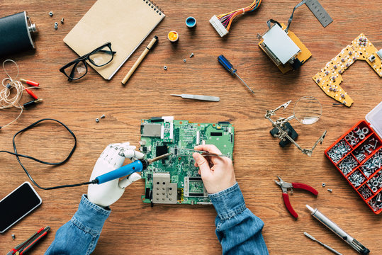 cropped image of electronic engineer with prosthetic arm fixing motherboard by soldering iron and tweezers