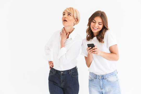 Portrait of caucasian woman 40s looking upward with disinterest while young teen girl holding and using black smartphone, standing isolated over white background