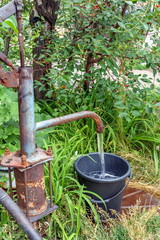 old rusty water column in the village fills the bucket with water on the background of dense grass and trees on a sunny day