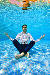 A young man with a beard in a white shirt sits underwater at the bottom of the pool in a Lotus position and meditates. Underwater photography. Vertical orientation