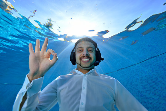Underwater portrait of a young man in a white shirt, with headphones on his head, who swims underwater in the pool, looking at the camera and smiling. Close up. Underwater photography