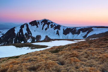 The meadow with the rocks and yellow grass. Landscape of high peaks of mountains covered with snow, pink sunrise at the horizon.
