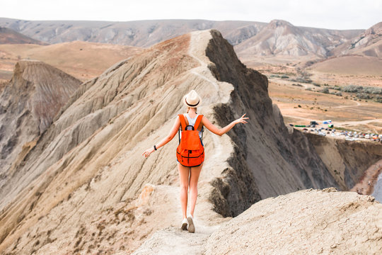 A girl in a straw hat and with a backpack walks along a mountain ridge in the middle of the desert.