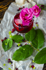 A glass with a red drink with berries, ice and a pink flower on crumpled wrapping paper beside which lie berries and a branch of green tropical leaves on a vintage wooden table