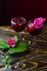 Two glasses full of red drink with berries, ice and a pink flowers, beside which lie berries and a branch of green tropical leaves on a vintage wooden table