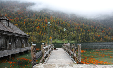Fototapeta premium An empty wooden pier by beautiful lakeside on a misty foggy morning ~ Gloomy autumn & Beautiful autumn scenery of Koenigssee (King's lake) in Bavaria, Germany