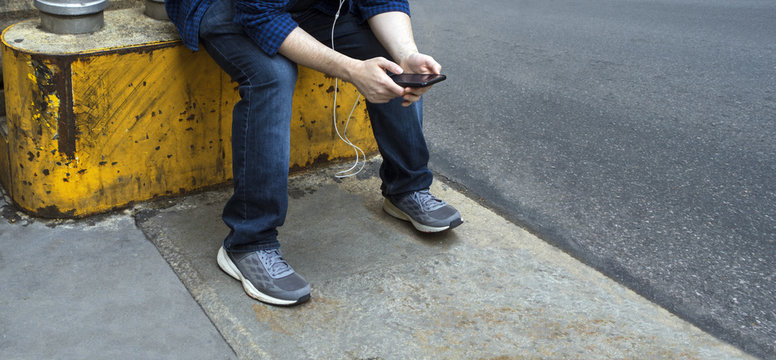 man with jeans and sneakers sitting on a yellow metal and cement blocker playing with his mobile device