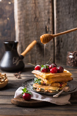 Freshly made belgian waffles with honey flows and powdered sugar. Cherries on top of waffles on wooden desk and napkin on wooden background. Coffee beans in glass jar. Turkish coffee pot for breakfast