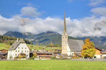 Fototapeta na wymiar A peaceful village at the foothills of Steinernes Meer Mountains in Maria Alm, Austria, with a majestic church tower (Wallfahrtskirche) standing tall in green fields & foggy mountains in background