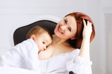 Portrait of a happy read-headed mother in a white dress breastfeeding her toddler boy. Mother care and relations concept
