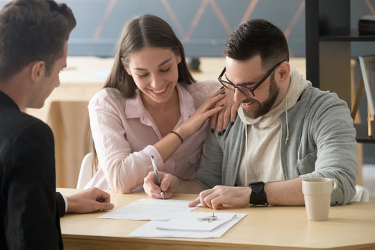 Excited millennial couple signing purchase agreement buying first home together, husband puts signature on document, becoming apartment owner, spouses legalize property ownership in realtor office