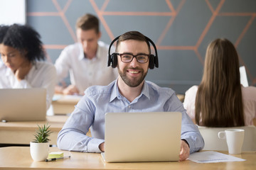 Portrait of happy male employee wearing headset looking at camera, working at laptop in coworking office, excited man posing for picture, smiling during corporate photoshoot in shared workspace