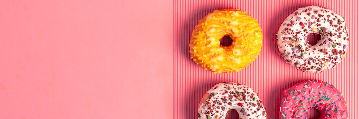 Panoramic view on four colorful donuts with sprinkles and icing on an empty pink pastel background
