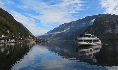 Fototapeta premium Autumn scenery of Hallstatt, a beautiful lakeside village in Salzkammergut area of Austria, with view of a tourist boat on the peaceful lake & reflections of mountains & the blue sunny sky on water
