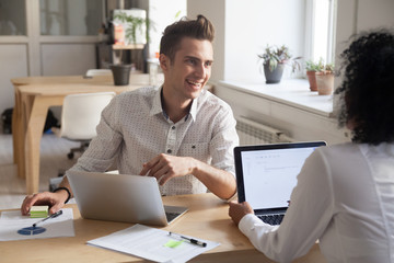 Smiling millennial colleagues talking while working at laptops, employees discussing business project or financial statistics in friendly office atmosphere, happy partners laughing during break