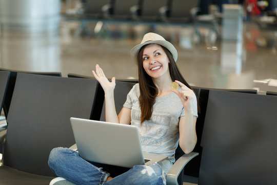 Young dreamy traveler tourist woman in hat sitting, working on laptop, holding bitcoin, spreading hands, waiting in lobby hall at airport. Passenger traveling abroad on weekend. Air flight concept.