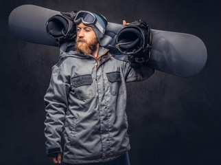Brutal redhead snowboarder with a full beard in a winter hat and protective glasses dressed in a snowboarding coat posing with snowboard at a studio, looking away. Isolated on gray background.