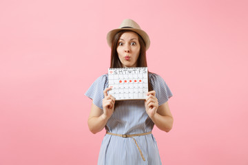 Portrait sceptical woman in blue dress, hat holding periods calendar for checking menstruation days isolated on bright trending pink background. Medical, healthcare, gynecological concept. Copy space.