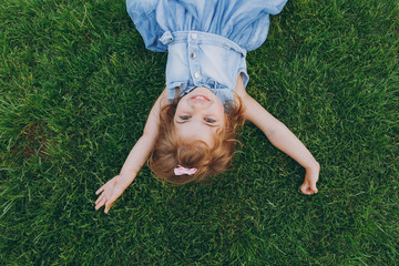 Joyful little pretty child baby girl in light dress lie on green grass lawn, spreading hands in...