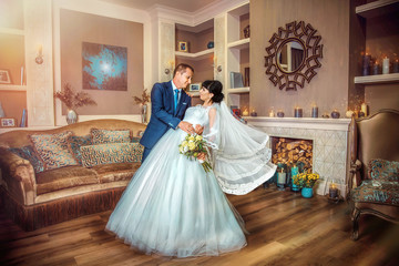 Groom gently tilting bride, holding her in his arms, on background of luxurious room with fireplace