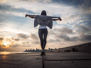 Healthy lifestyle: A happy athlete walks after jogging and spread her arms in the sides against the backdrop of the city and sunset.