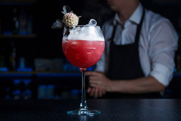 Pink, chilled cocktail in a glass for red wine with gin and wine syrup  is decorated with a flower, stands on the bar counter with a barmen in the background. Сocktail card, cocktail menu