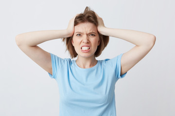 Portrait of angry irritated young woman in blue tshirt standing with hands on head and feels crazy isolated over white background Looks directly in camera