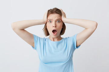 Closeup of amazed shocked young woman in blue t shirt standing with opened mouth and hands on her head isolated over white background Feels stunned and worried