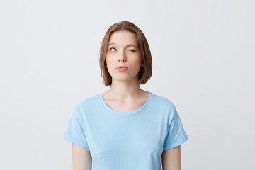 Fototapeta premium Thoughtful concentrated young woman in blue t shirt with one closed eyes thinking and trying to remember answers for the questions on exam isolated over white wall Looking to the side and up