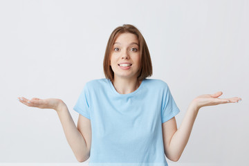 Portrait of confused cute young woman in blue t shirt shrug her shoulders and hold copyspace on both palms isolated over white background