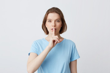 Portrait of peaceful beautiful young woman in blue t shirt looks directly in camera and showing silence gesture isolated over white background