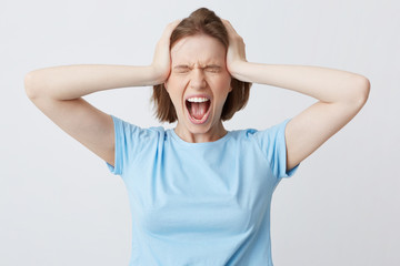 Portrait of crazy hysterical young woman in blue tshirt with closed eyes and hands on head looks stressed and screaming isolated over white background