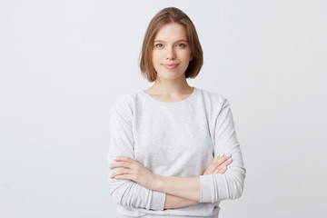 Fototapeta premium Zbliżenie szczęśliwa ładna młoda kobieta w longsleeve stojącej z rękami splecionymi i patrząc bezpośrednio w kamerę na białym tle nad białą ścianą Czując się pewnie