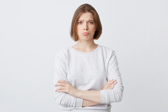 Closeup Of Sad Unhappy Pretty Young Woman In Longsleeve Stands With Hands Folded And Feels Displeased Isolated Over White Background Looks Directly In Camera