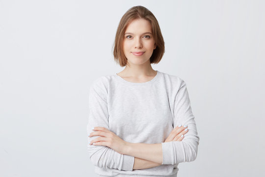 Closeup of happy pretty young woman in longsleeve standing with hands folded and looking directly in camera isolated over white wall Feeling confident