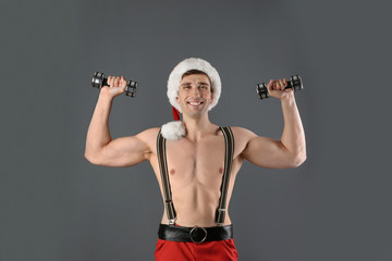 Fototapeta na wymiar Young muscular man in Santa hat with dumbbells on gray background
