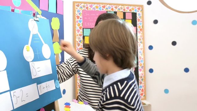 Cute boy solving graphic math exercises at kindergarten, under educator observation